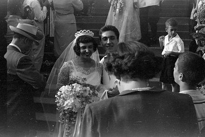 A black and white photograph of a bride wearing a tiara, veil, and lace dress, holding a bouquet of flowers. She is standing ...