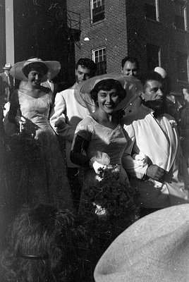 A black and white photograph of a group of people standing outdoors against a brick building. Two women wear wide-brimmed hat...