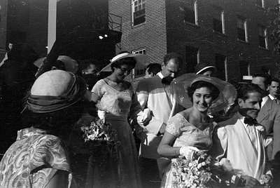 A black and white photograph of a group of people in formal attire standing outdoors against a brick building. Two women wear...