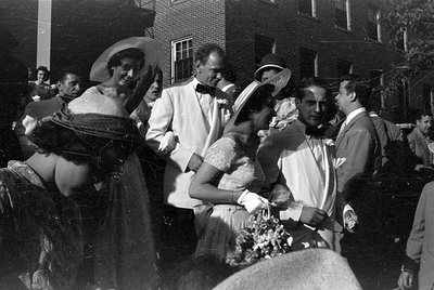 A group of people in formal attire gathered outdoors in front of a brick building. Several men wear white dinner jackets with...