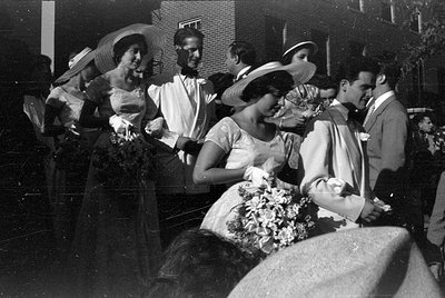 A group of people in formal attire stand outdoors in front of a brick building. Several women wear wide-brimmed hats and glov...