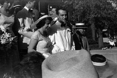 A black and white photograph of a group of people in formal attire outdoors. A woman in a light-colored dress and wide-brimme...