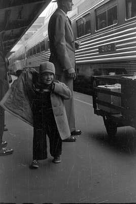 A black and white photograph shows a young child in a heavy coat and cap standing on a platform next to a corrugated metal tr...