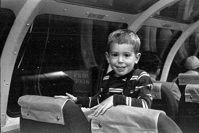 A young boy with short hair and a striped sweater sits inside a vehicle with large, curved glass windows. He is looking towar...