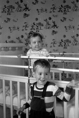 A black and white photograph of two young children. One child stands in the foreground wearing a striped long-sleeve shirt an...