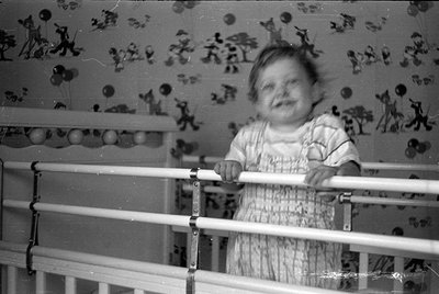 A black and white photograph of a young child standing in a white metal crib. The child wears a light-colored patterned dress...