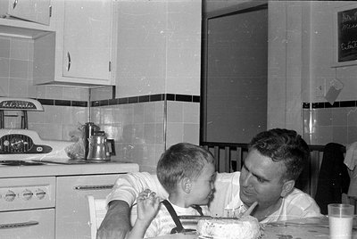 A man and a young boy in a kitchen. The man is leaning toward the boy, and a frosted cake sits on the table in the foreground...