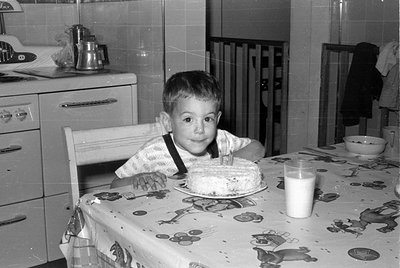 A young boy sits at a table covered with a patterned tablecloth, looking toward the camera. In front of him is a frosted cake...