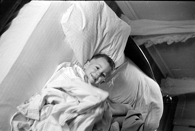 A black and white photograph of a young child lying in a bed. The child is tucked under a striped blanket and resting their h...