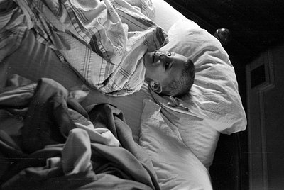 A black and white photograph of a baby lying on a white pillow. The child is partially covered by a striped blanket and a dar...