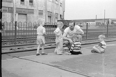 A man in a white t-shirt and three young children are gathered on a paved area next to railway tracks. In the background is a...