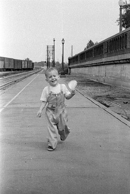 A young child wearing light-colored overalls and a t-shirt runs along a concrete train platform. A line of freight cars is vi...