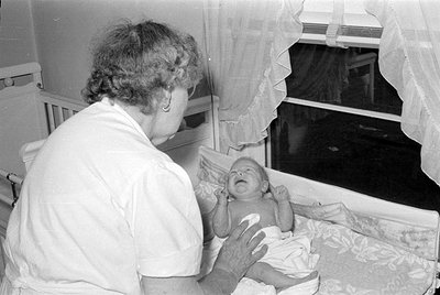 A black and white photograph showing a woman from behind, wearing a white short-sleeved uniform, looking at a crying infant i...