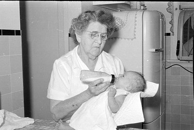 A black and white photograph of an older woman with glasses feeding a baby from a bottle. The baby is wrapped in a white clot...