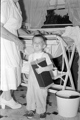 A black and white photograph of a young child in striped overalls holding a large rectangular box. A woman in a white dress s...