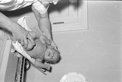 A black and white photograph showing a baby lying on a medical scale. A person's hands are visible, holding the baby in place...
