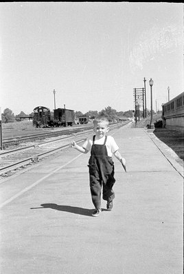 A young boy wearing dark overalls and a white t-shirt walks along a concrete train platform. In the background, several railc...