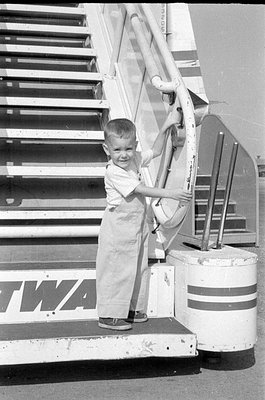 A young child wearing light-colored overalls stands on the bottom step of a mobile aircraft boarding staircase. The letters '...