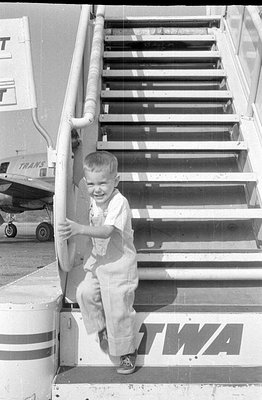 A black and white photograph showing a young child in light-colored clothing leaning over a railing. In the background, a str...