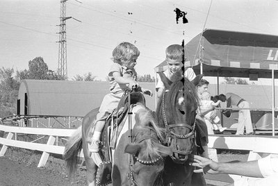 A black and white photograph of two young children sitting on horses. In the background, a corrugated metal building, a tall ...