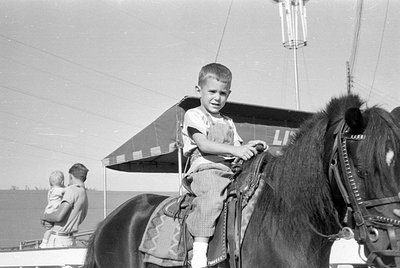 A young boy in overalls sits atop a dark-colored horse with a decorative saddle. In the background, a man holds a toddler, an...