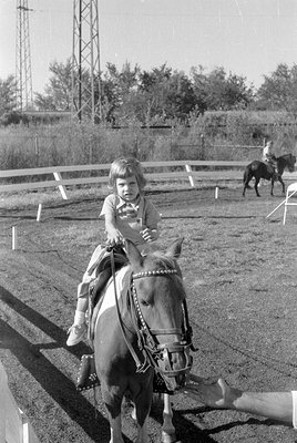 A young child sits atop a pony in an outdoor riding arena. The pony wears a decorative bridle with metallic studs. In the bac...