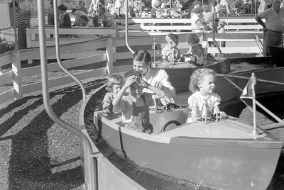 Black and white photograph of children in a boat ride at an amusement park. Three children sit in a small boat in the foregro...