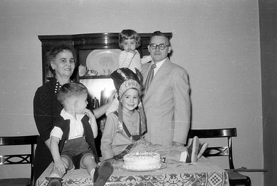 A black and white indoor photograph of a woman, a man in a suit and glasses, and three children. One child wears a Native Ame...