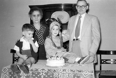 A black and white photograph of a man, woman, and two children gathered around a table with a frosted cake. One child wears a...