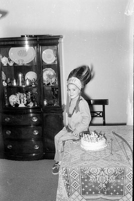 A child wearing a Native American-style headdress and costume sits at a table with a frosted cake. In the background is a dar...