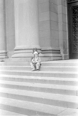 A child wearing a feathered headdress and light-colored clothing sits on wide stone steps. Behind the child are large, smooth...