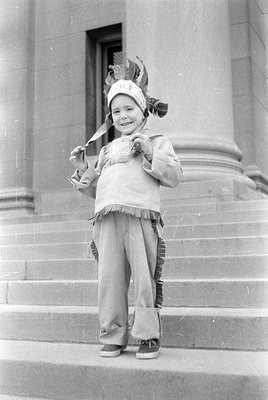 A young child in a Native American-inspired costume stands on stone steps outside a building with columns. The outfit include...