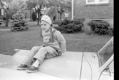Child in 1960s-style Native American headdress sits on a car hood in a residential driveway; overalls, knee-high socks, and s...