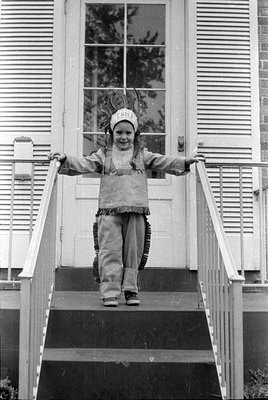 Child in 1950s–60s Native American-themed costume (chief headdress, fringed vest) posing on front stairs of a residential hou...