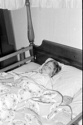 A person lies on a bed with floral-patterned bedding, partially covered by a blanket. The bed has a wooden headboard with a c...