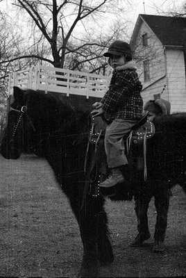 A person in winter gear rides a horse on a paved path beside a white fence and a house. The rider wears a cap, plaid shirt, a...