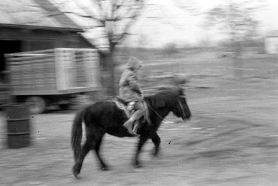 Blurred black-and-white photo of a person riding a horse on a paved road, with motion blur obscuring details. A truck and bui...