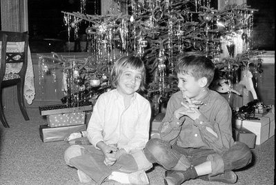 Two children sitting on the floor in front of a decorated Christmas tree with wrapped gifts, smiling at the camera. Indoor ho...