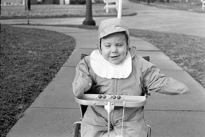 Child in a light-colored, hooded raincoat riding a small pedal cart on a paved sidewalk near grass. The child appears to be s...