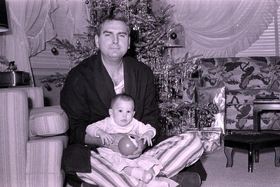 Black-and-white indoor portrait of an adult holding a baby in front of a decorated Christmas tree, likely mid-20th century. T...