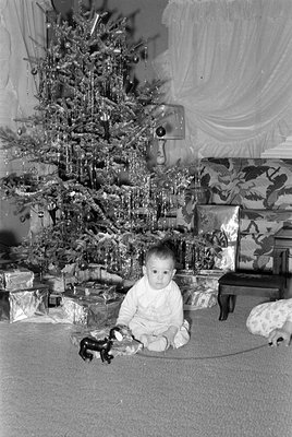 A young child sits on a carpeted floor in front of a decorated Christmas tree with wrapped gifts, holding a small stuffed ani...