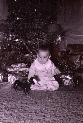 A young child in a white onesie sits on a textured floor beside a small black-and-white toy dog, surrounded by wrapped presen...