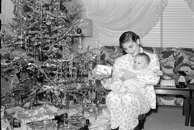 A woman in a floral robe holds a baby while standing near a decorated Christmas tree with wrapped gifts beneath it. Indoor se...