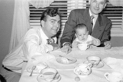 Three men and a baby seated at a table with tea cups, saucers, and food; indoor setting with striped curtains. Two men in sui...