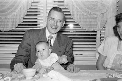 A man in a suit holds a baby at a table with a woman in a dress partially visible; indoor setting with striped curtains and t...