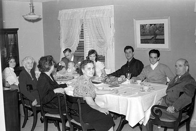 Black-and-white indoor group portrait of nine adults seated around a table set for a meal, likely a holiday dinner. A woman i...