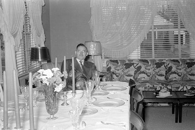 A man in a suit sits at a formal dining table set with glassware, plates, and a floral centerpiece. Decorative curtains and p...