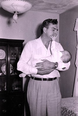 A man in a light-colored shirt and trousers holds a swaddled infant indoors, near a glass-fronted cabinet with glassware. Cei...