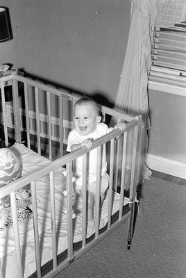 Black-and-white photo of an infant standing in a crib, smiling, wearing a diaper and short-sleeved shirt. A stuffed animal an...