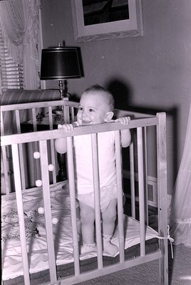 Infant standing in a wooden crib, smiling at the camera. Indoor setting with a lamp, patterned bedding, and a vent visible. M...
